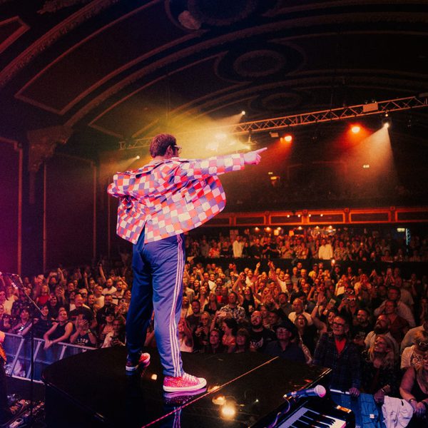 A man dressed as Elton John performing on stage. He is facing away from the camera towards a crowd.