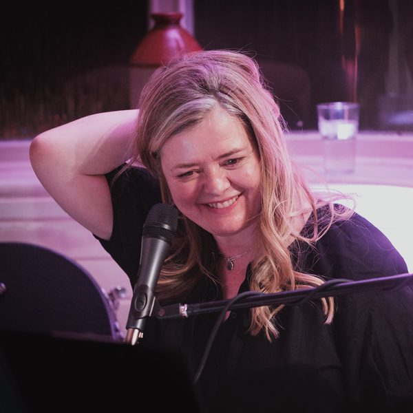 A woman on stage in front of a microphone. She is smiling with her hand in her hair.