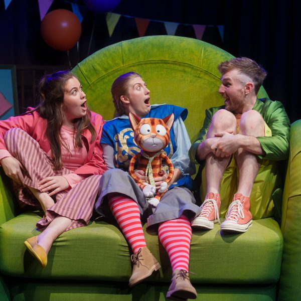 The cast of Sir Charlie Stinky Socks performing onstage, sat in colourful outfits on a large green couch.
