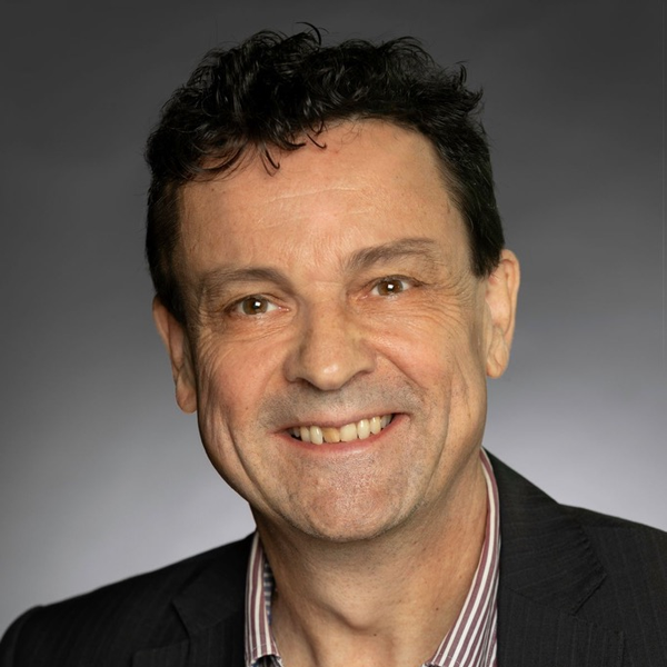 Studio headshot of Damien Lewis, wearing a dark blazer over a striped shirt, smiling at the camera against a plain grey background, with two lapel pins visible on the jacket.