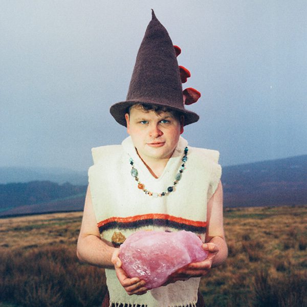 Comedian Dan Tiernan stood in front of a sweeping flat landscape. He is wearing a witch's hat, a white shirt, a crystal necklace and is holding a large piece of rose quartz.