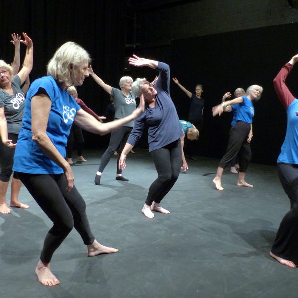 A group of older adults performing expressive dance movements on a theatre stage under stage lighting.