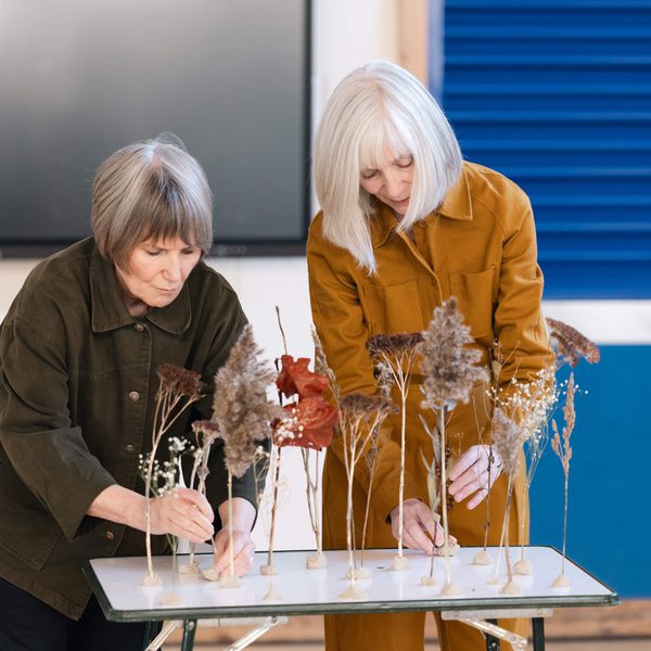 Two women putting flower stems into position on a small white table.