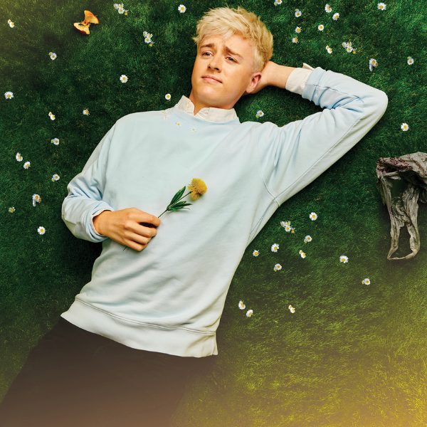 Person lying on grass dotted with daisies, wearing a pale blue sweatshirt and holding a dandelion flower.