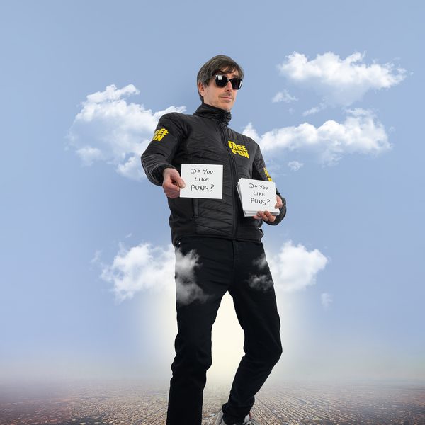 Darren Walsh standing on a cityscape in front of a blue, cloudy sky, as if a giant, holding cards with the handwritten phrase "Do you like puns?"