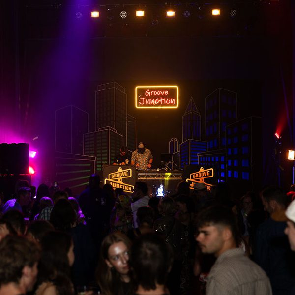 A DJ playing in front of a crowd, with a neon cityscape and a sign that says 'Groove Junction' behind him.