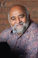 A head shot of East is East cast member Bhasker Patel. He has a bald head and beard and wears a paisley shirt.