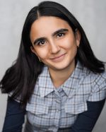 A headshot of East is East cast member Maya Dhokia. She has long hair and wears a buttoned-up shirt over a long-sleeved top.