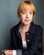 A headshot of East is East cast member Victoria Brazier. Her long hair is tied back. She's wearing a denim jacket and a watch, with one arm crossed in front of her chest.