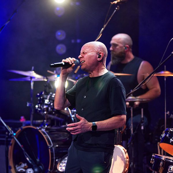 A band performing on stage, with the lead singer dressed similarly to Phil Collins and singing into a microphone.