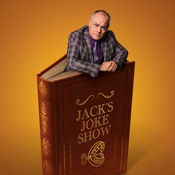 Jack Dee leaning out from the top of a giant hardback book against a warm orange background. The book is dark red with gold decorative detailing and the title “JACK’S JOKE SHOW” on the cover, along with an illustration of a wind-up chattering teeth toy.