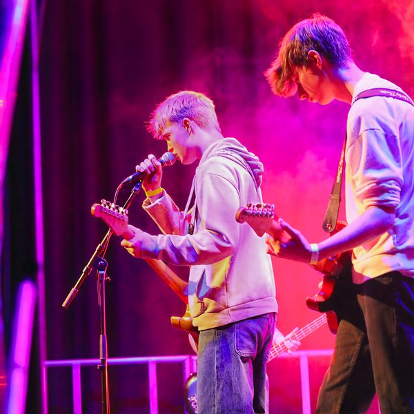 Two teenage boys playing guitar and singing on stage, washed in pink light.