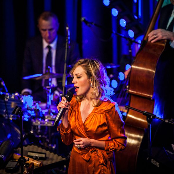 A woman in an orange dress singing into a microphone on stage. Several musicians, including a double bassist, are behind her.