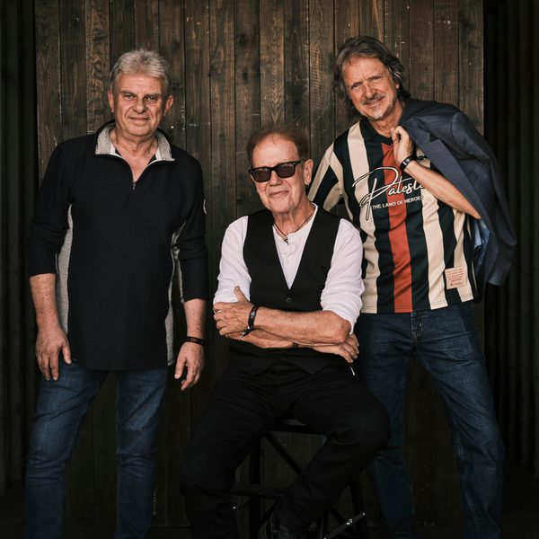 John Jones, Ray Cooper and Al Scott of the John Jones Trio pose in front of a wooden backdrop, with one seated in the centre and two standing on either side.