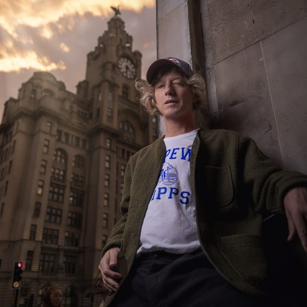 John Power wearing a green jacket and baseball cap leaning against the corner of a building with Liverpool's Liver building and a sunset sky in the background.