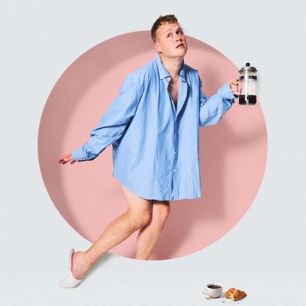 Josh Jones wearing an oversized light blue button-down shirt balances on one foot against a pale grey studio backdrop with a large pink circle, Holding a coffee pot in one hand, while a small cup of coffee and a croissant sit on the floor nearby