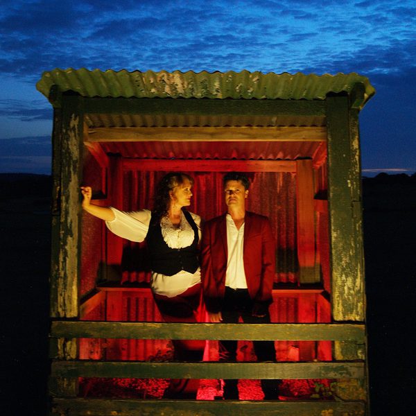 Two people stand side by side inside a small, weathered wooden shelter with a corrugated roof, illuminated by red light from below, in front of a deep blue evening sky.