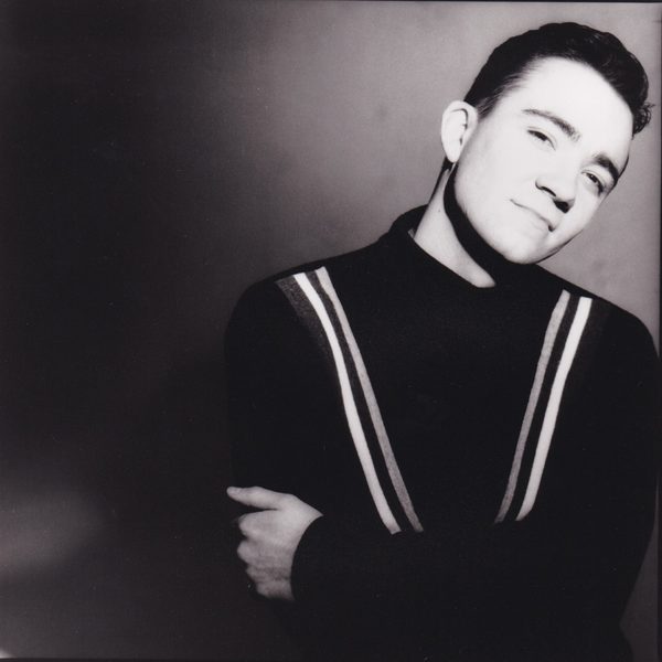 Black-and-white portrait of Kenny Thomas in a dark jumper with light shoulder stripes, arms crossed and head tilted, against a dark background