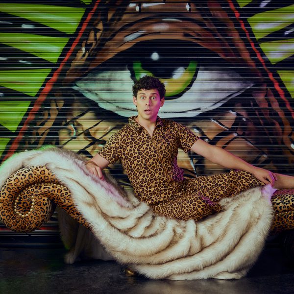 Matt Richardson wearing a leopard-print outfit reclines on an ornate chaise lounge covered in white faux fur and leopard-print fabric. Behind the chaise is a large, colourful mural painted on a metal shutter, featuring a single green eye centred within a triangular design.