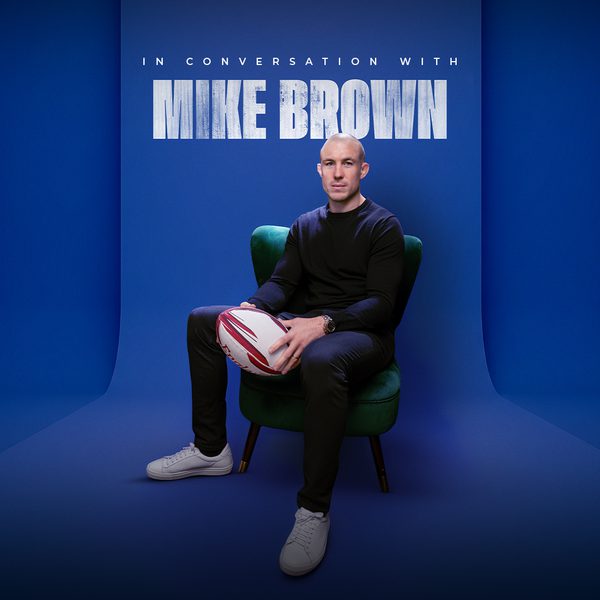 Rugby player Mike Brown sitting and holding a rugby ball against a blue background. Text: In Conversation with Mike Brown.