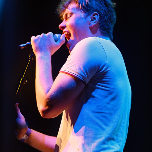 A young person in a white t-shirt singing into a microphone on stage.
