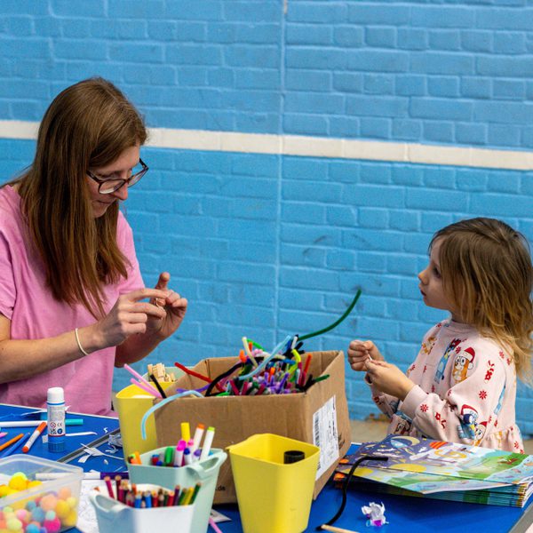 A woman and a child sat facing each other in front of a blue wall. They are making crafts from pipe cleaners, and a pile of other supplies is on the table in front of them.