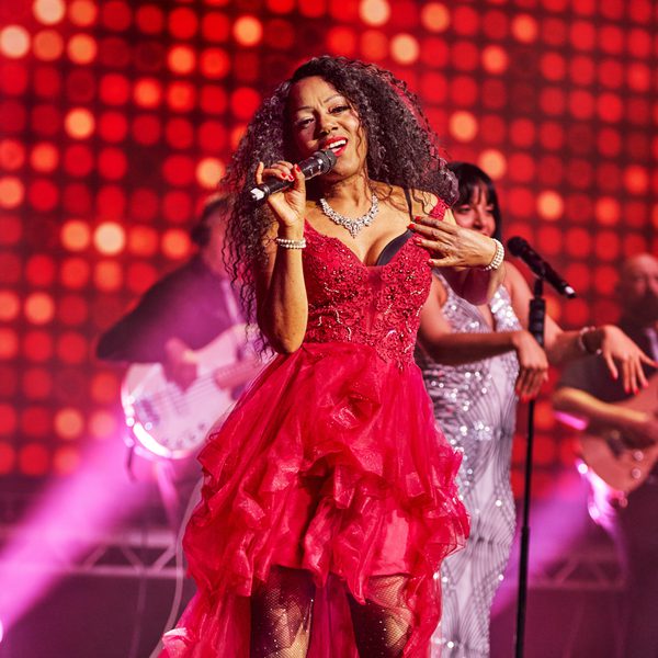 A woman in a sparkly red dress singing into a microphone on stage against a lit up red backdrop, with two backing singers.