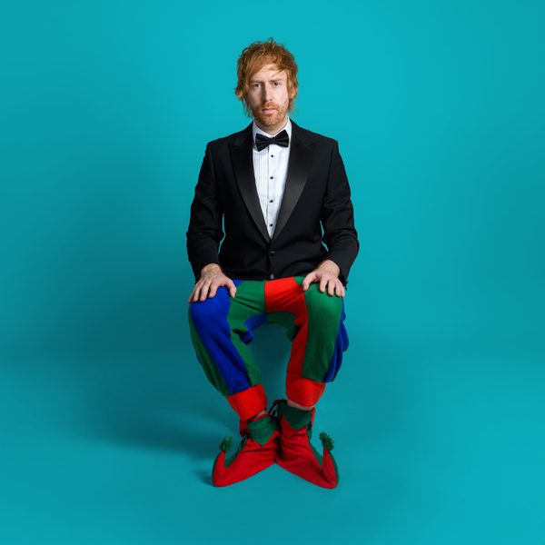 Comedian Mark Simmons in a tuxedo jacket and jester trousers, sat on a stool against a blue background.