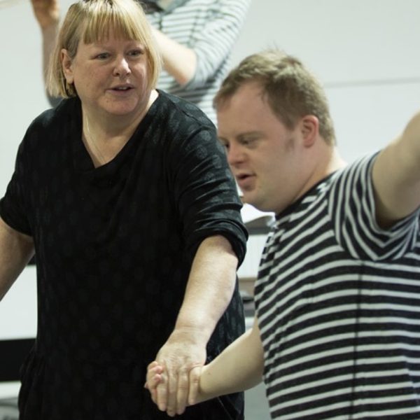 Nathan Bessell and Heather Williams in a rehearsal room standing side by side, holding hands. Heather is in a black top and faces Nathan, who is wearing a striped shirt and extends an arm and points toward the right side of the room.