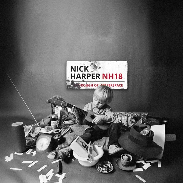 Black and white: A small boy playing a child-size guitar surrounded by a pile of miscellaneous objects. A street sign is behind him, reading: Nick Harper NH18, Borough of Happerspace.