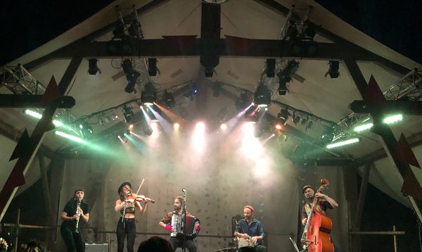 A band performs on a large stage beneath a triangular tent roof. Multiple musicians play clarinet, violin, accordion, percussion, and double bass. The audience is visible in silhouette in the foreground, with stage lights overhead.