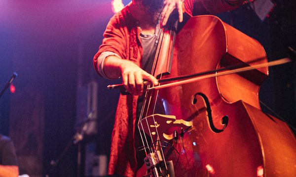 Close-up of a musician playing a double bass on stage under pink and purple lighting. The musician uses a bow on the strings, with microphones and cables attached to the instrument. Stage lights and other performers are partially visible in the background