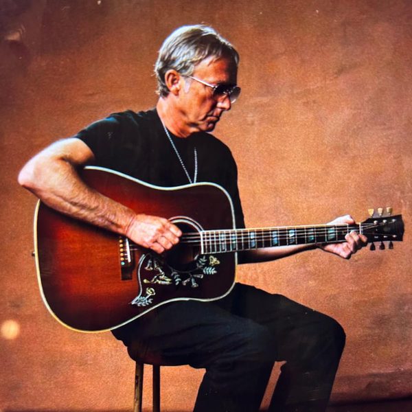 Guitarist Steve Cradock dressed in all black, sat on a stool against a brown background strumming a guitar.