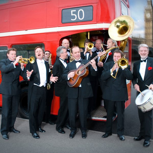 The members of the Pasadena Roof Orchestra dressed in tuxedos and holding various instruments in front of a red London bus.