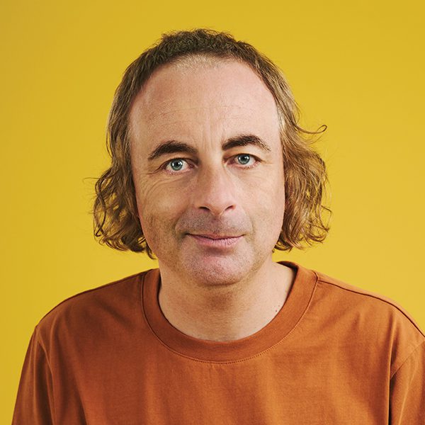 Headshot of Paul Foot, with shoulder-length wavy hair wearing a rust-coloured T-shirt, facing the camera against a solid bright yellow background.
