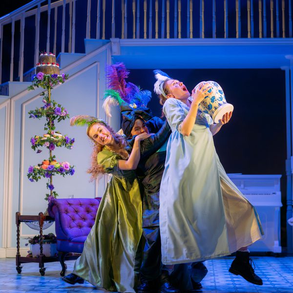 Three actresses in regency costumes performing on stage. They are struggling with each other for a large vase, and are standing in front of a chaise and a floral cake tower.