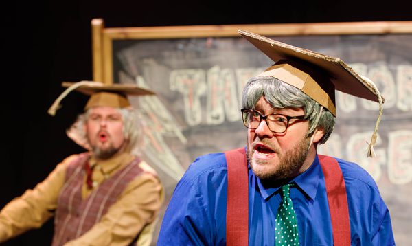 Two men dressed as professors in cardboard hats performing on stage in front of a large blackboard.