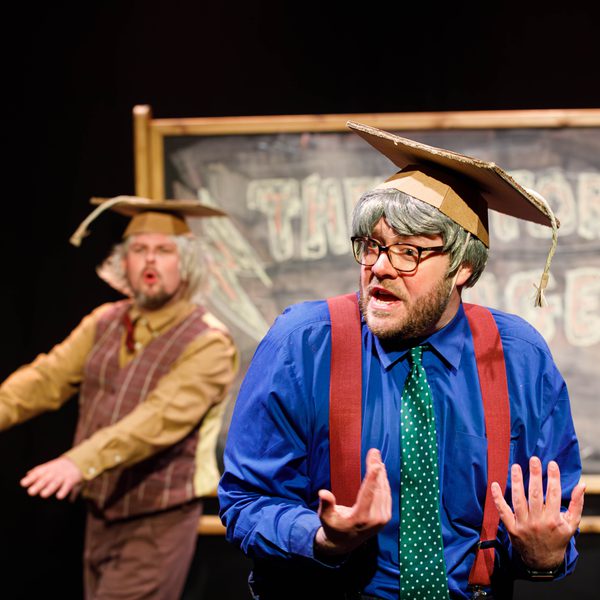 Two men dressed as professors in cardboard hats performing on stage in front of a large blackboard.