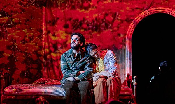 The cast of The Beekeeper of Aleppo performing on stage. A man and a woman sit on a mound of rubble, while red projections encompass the stage.