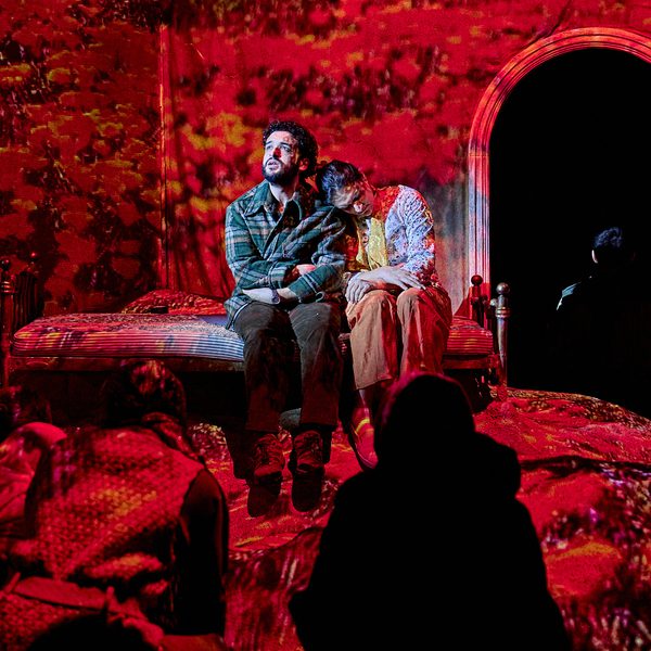 The cast of The Beekeeper of Aleppo performing on stage. A man and a woman sit on a mound of rubble, while red projections encompass the stage.