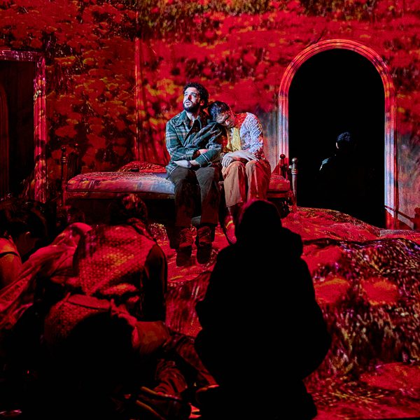 The cast of The Beekeeper of Aleppo performing on stage. A man and a woman sit on a mound of rubble, while red projections encompass the stage.