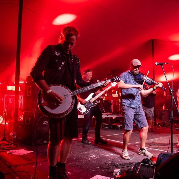 Two musicians performing on an outdoor stage, one playing a banjo and the other playing a violin. They are washed in red light.