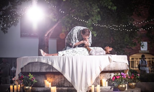 Will Fletcher and Grace Wylde as Romeo and Juliet on top of a round stone fountain dressed to look like a tombstone. Juliet appears to be dead and Romeo is kissing her hand.