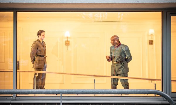 Patrick Fusco as Paris and Mark Springer as Lord Capulet stand inside a brightly lit corridor, viewed through a large floor-to-ceiling window from outside. The room has warm yellow lighting, smooth walls and hanging wall lamps. A railing runs along the outside in front of the window.