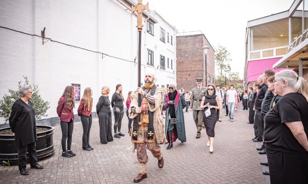 A group of people stand in two facing lines in a narrow outdoor courtyard between buildings while a central figure in ornate, historical-style costume walks down the middle; the setting includes brick walls, white building façades, and a small tree in a planter.