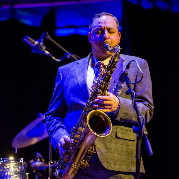 A man playing the saxophone on stage, dressed in a grey suit.