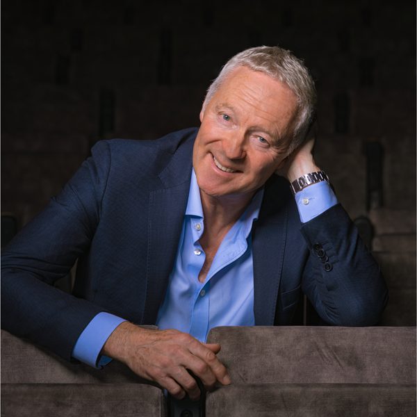 Impressionist Rory Bremnar, smiling in a blue suit and leaning over a row of grey velvet seats.
