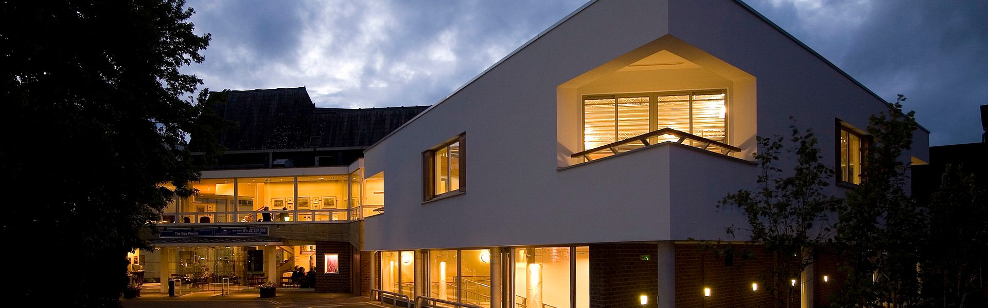 Salisbury Playhouse at dusk, windows glowing with warm yellow light. Trees frame the scene, and a cloudy blue-grey evening sky fills the background.