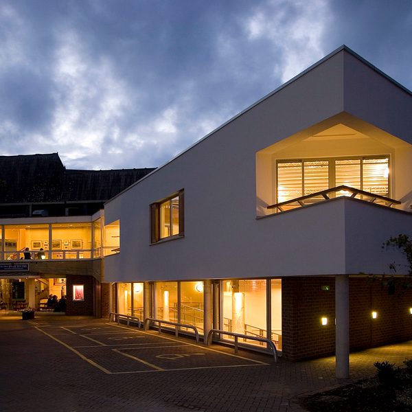 Salisbury Playhouse at dusk, windows glowing with warm yellow light. Trees frame the scene, and a cloudy blue-grey evening sky fills the background.