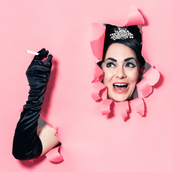 A woman dressed as Audrey Hepburn bursting through a pink background, smiling and holding a cigarette.